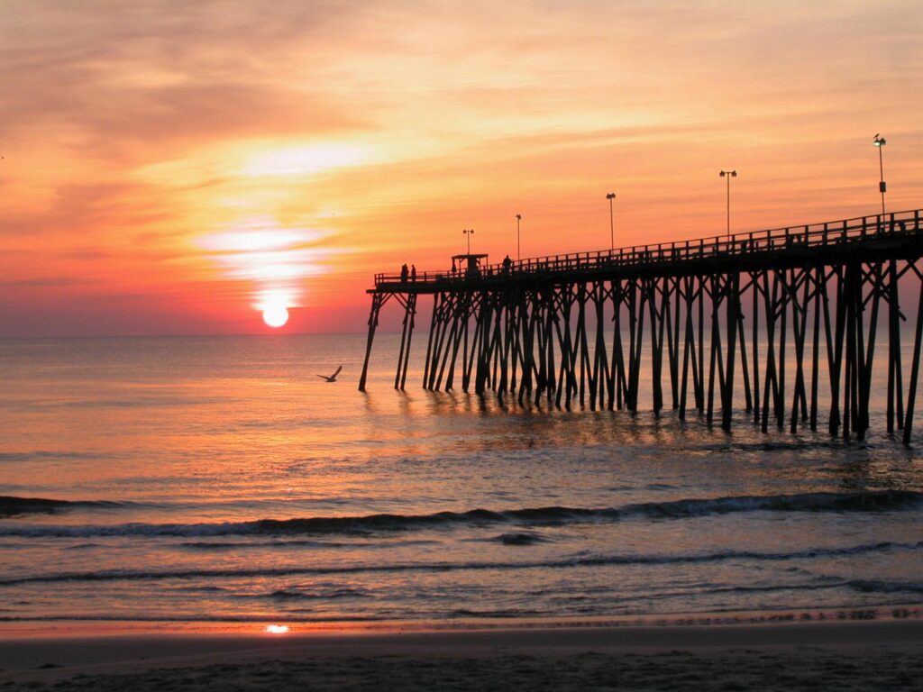 The Kure Beach Fishing Pier silhouetted against a sunrise over the Atlantic, with a gull and breaking waves in the foreground