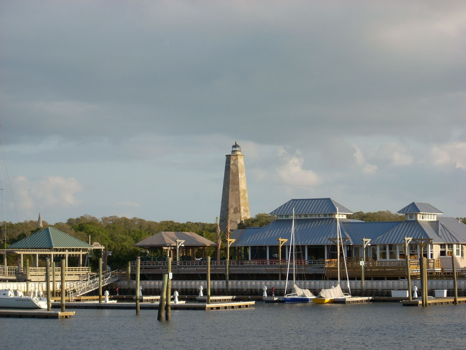 Bald Head Island Marina on Bald Head Island, North Carolina, with sailboats at the docks, the Harbour Village buildings along the waterfront and the Old Baldy lighthouse rising above the maritime forest behind them