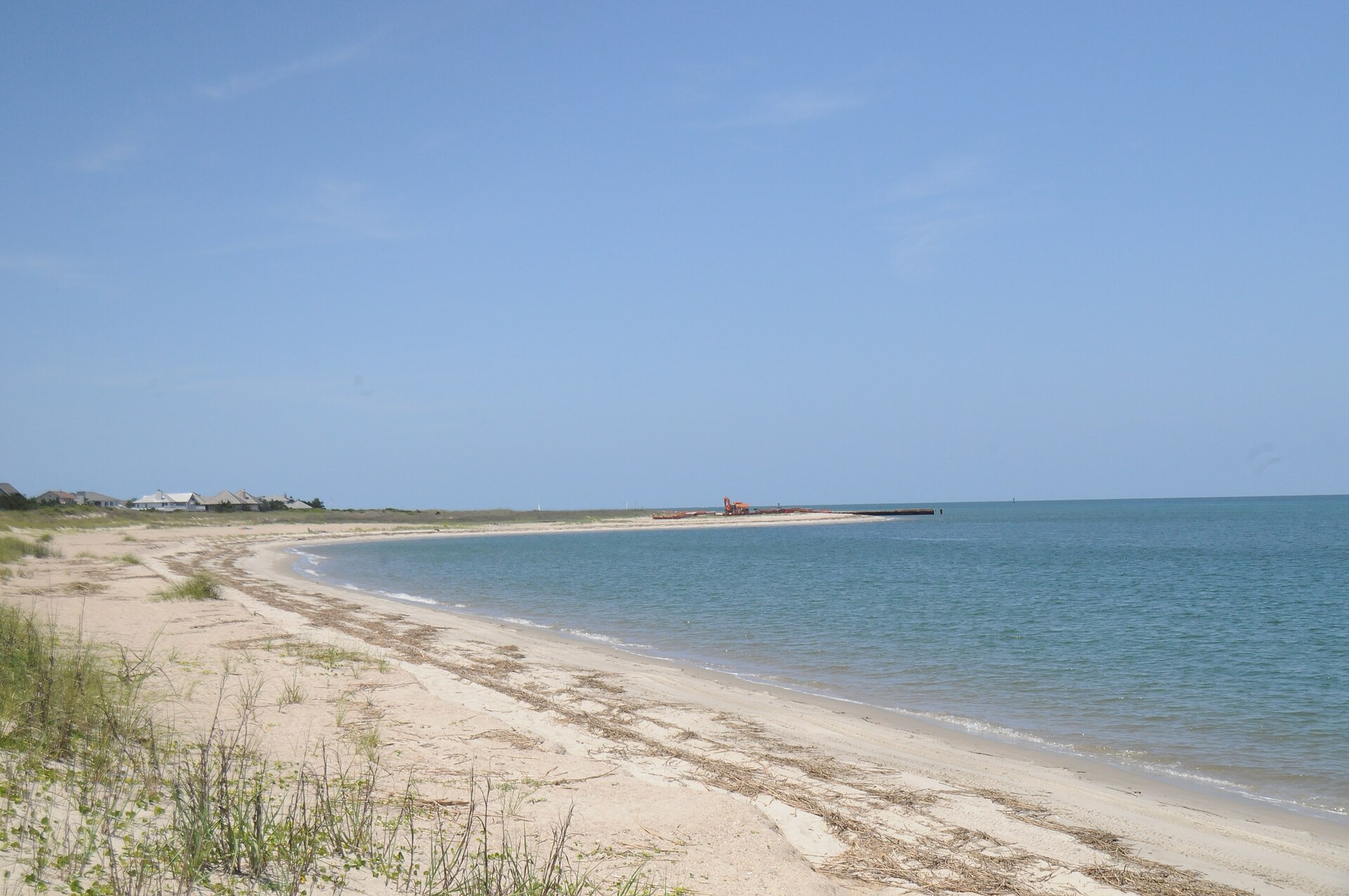 The curving sand beach on the Cape Fear River side of Bald Head Island, North Carolina, with dune grass in the foreground, a row of houses set back behind the dune line and the point in the distance
