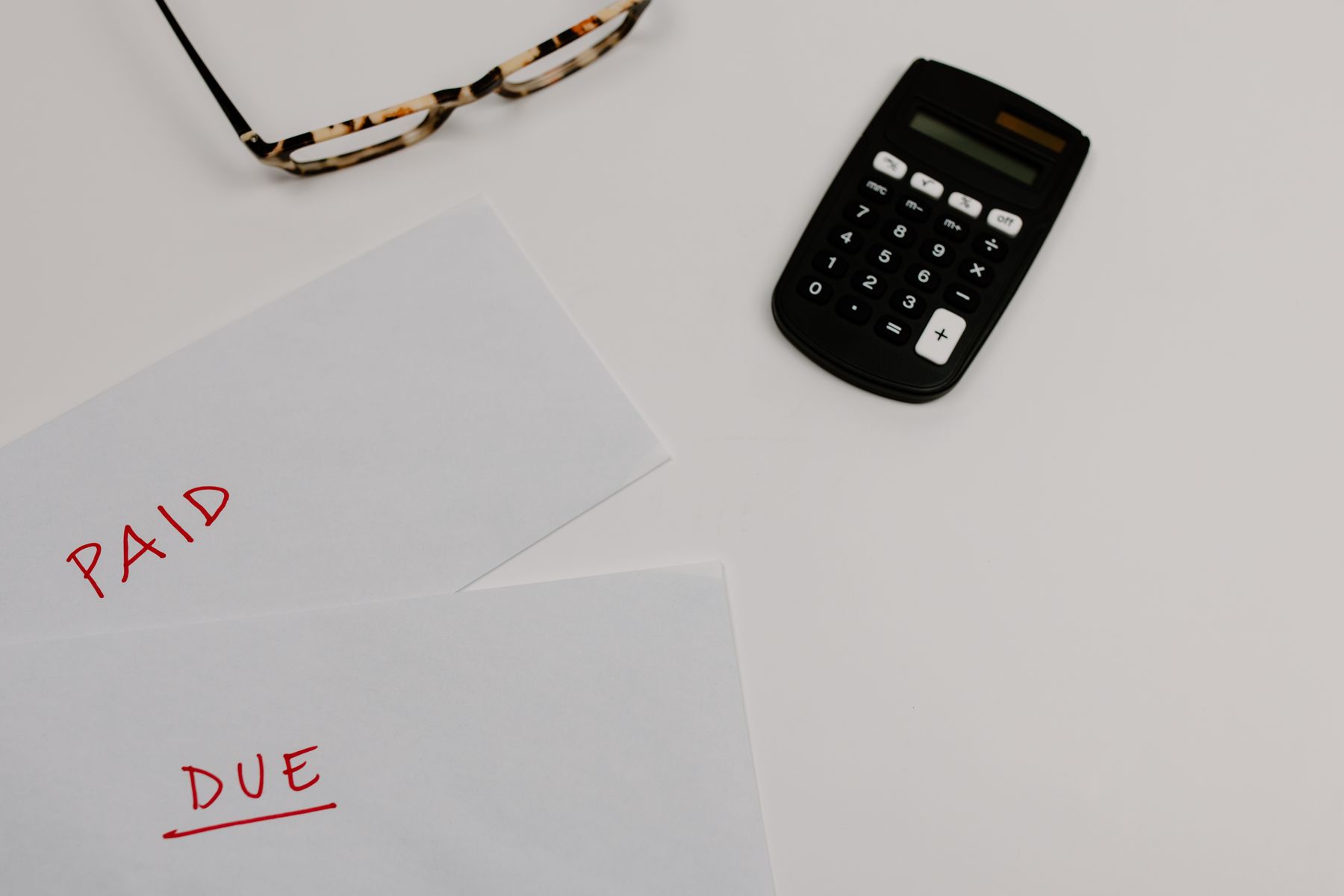 Household bills marked paid and due on a desk beside a calculator and reading glasses
