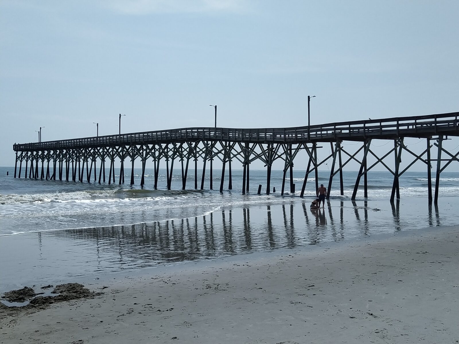 The Holden Beach Fishing Pier reaching out over the surf at Holden Beach, North Carolina, with the pier pilings reflected in the wet sand at low tide