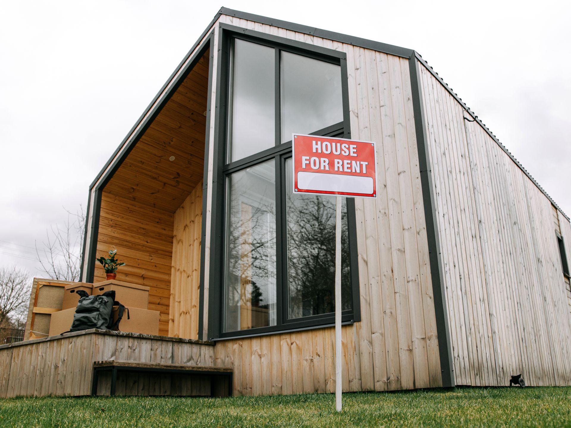A red House For Rent sign on the lawn in front of a wood-clad rental home, with moving boxes stacked on the front step