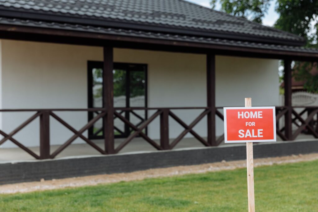 A red Home for Sale sign in the front yard of a house with a covered porch
