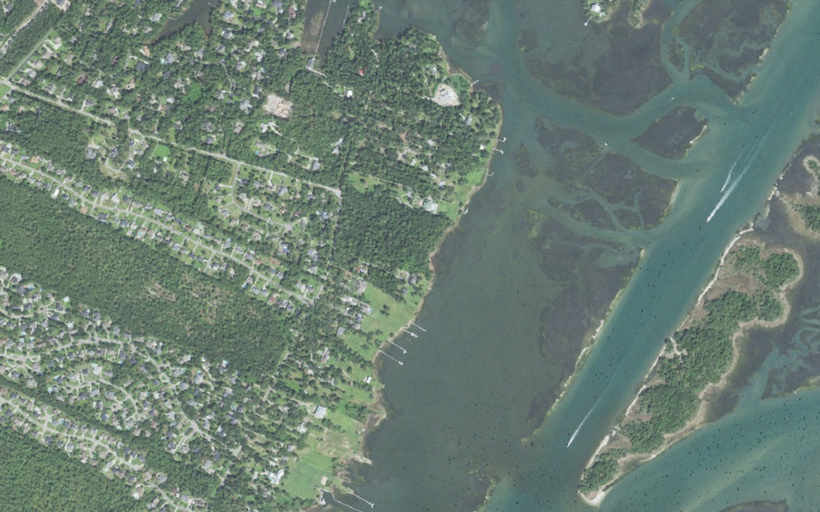 Aerial view of the sound side of Masonboro in New Hanover County, North Carolina, showing wooded neighborhoods off Masonboro Sound Road, private docks reaching across the marsh to the Intracoastal Waterway, and the undeveloped Masonboro Island Reserve beyond the channel