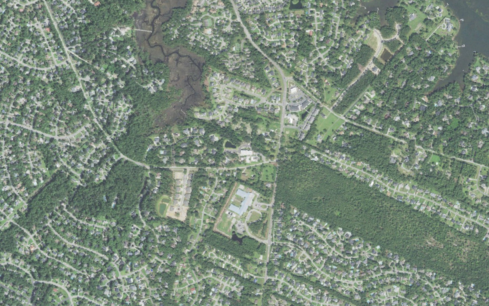 Aerial view of the Masonboro Loop Road corridor in Wilmington, North Carolina, with Masonboro Elementary School and the shops at the Masonboro Loop and Pine Grove crossroads surrounded by wooded subdivisions and the Whiskey Creek marsh