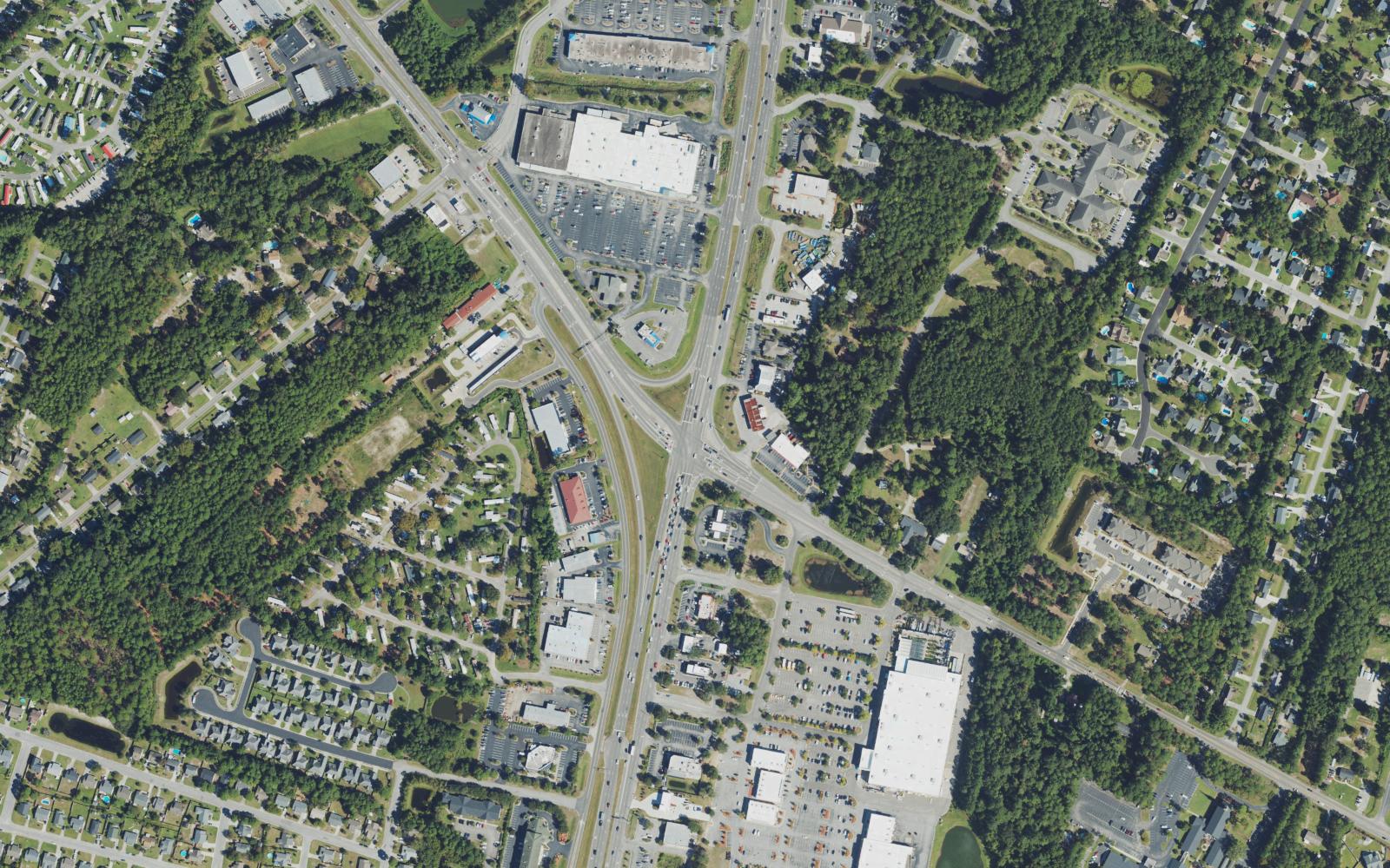 Aerial view of the Monkey Junction intersection in southern New Hanover County, North Carolina, where US 421 Carolina Beach Road and NC 132 South College Road converge, showing the big box stores and shopping center parking lots on all four corners and the wooded subdivisions and townhome rows around them