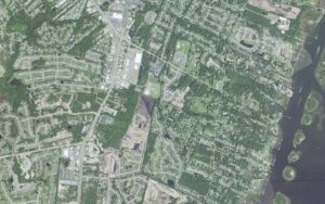 Aerial view of the heart of Myrtle Grove in New Hanover County, North Carolina, showing the Carolina Beach Road commercial corridor running diagonally through the frame, wooded subdivisions under heavy tree canopy on both sides, the Myrtle Grove Athletic Complex ball fields, and the Intracoastal Waterway with private docks along Masonboro Sound at right