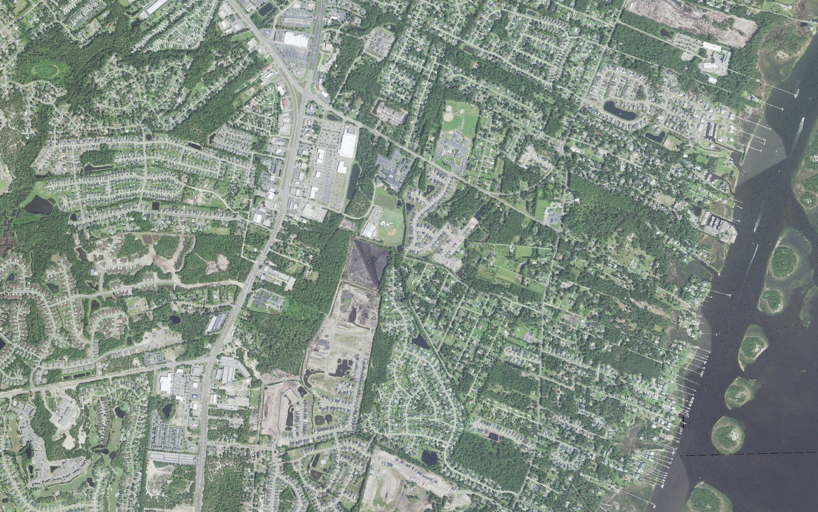 Aerial view of the heart of Myrtle Grove in New Hanover County, North Carolina, showing the Carolina Beach Road commercial corridor running diagonally through the frame, wooded subdivisions under heavy tree canopy on both sides, the Myrtle Grove Athletic Complex ball fields, and the Intracoastal Waterway with private docks along Masonboro Sound at right