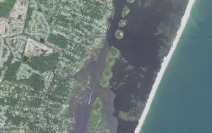 Aerial view of the sound side of Myrtle Grove, North Carolina, with rows of private docks reaching from Masonboro Loop Road across the marsh to the Intracoastal Waterway channel, the undeveloped Masonboro Island Reserve beyond it, and the Atlantic Ocean at right
