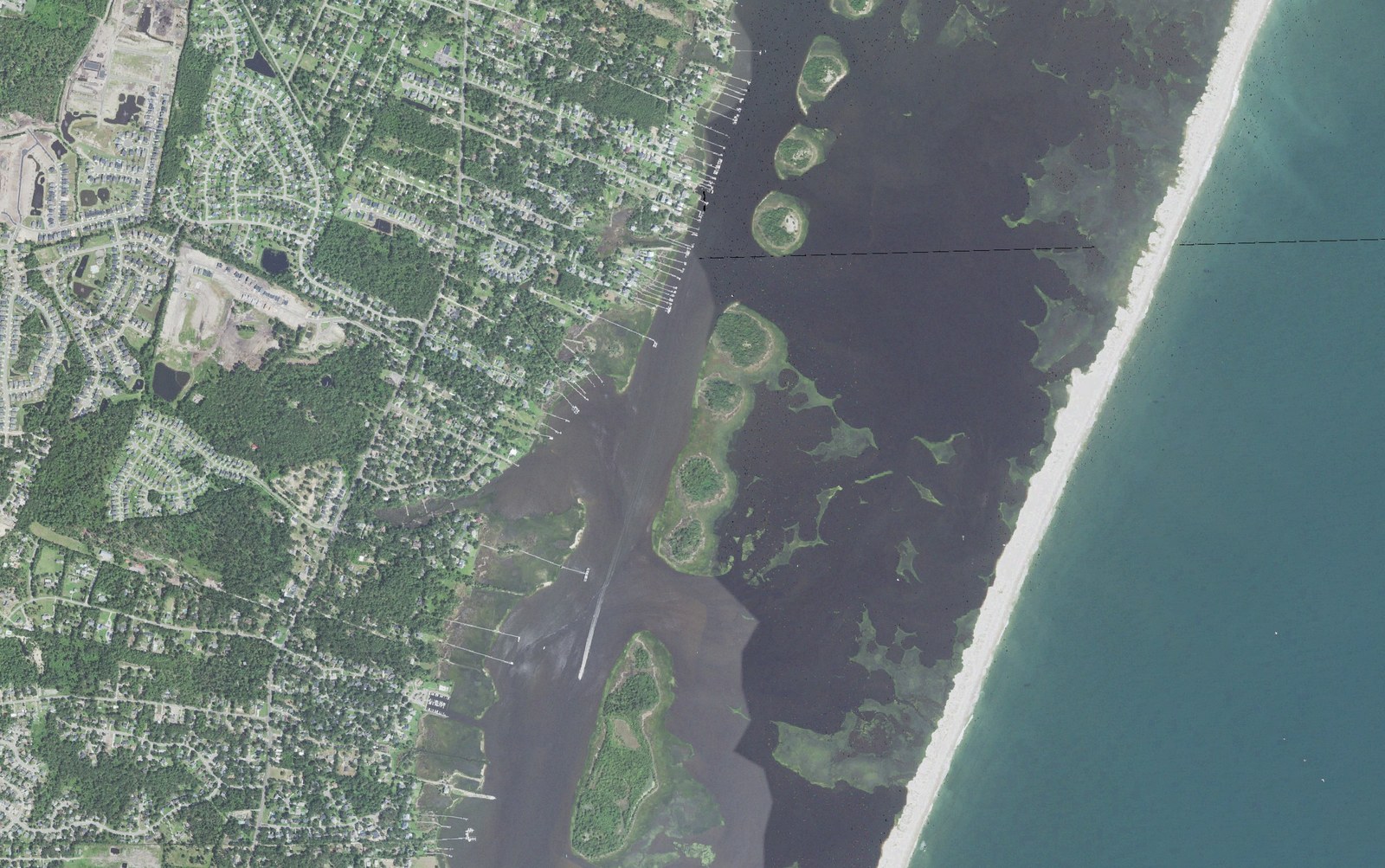 Aerial view of the sound side of Myrtle Grove, North Carolina, with rows of private docks reaching from Masonboro Loop Road across the marsh to the Intracoastal Waterway channel, the undeveloped Masonboro Island Reserve beyond it, and the Atlantic Ocean at right