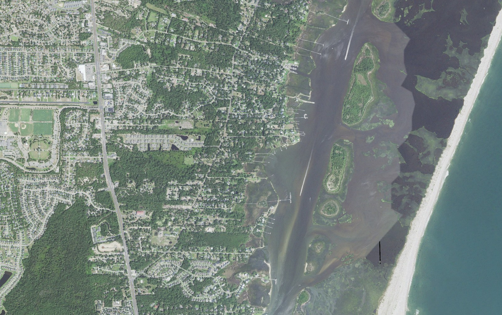 Aerial view of southern Myrtle Grove, North Carolina, showing Carolina Beach Road and its ball fields at left, newer subdivisions and wooded lots through the center, waterfront homes with docks along Myrtle Grove Sound, and Masonboro Island with the Atlantic beach at right