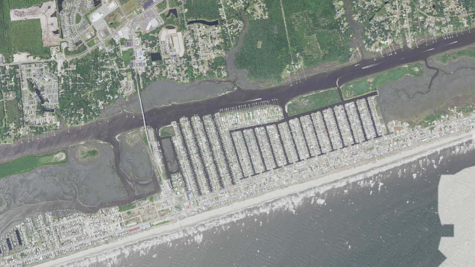 Aerial view of the center of Ocean Isle Beach, North Carolina, showing the Causeway Drive bridge crossing the Intracoastal Waterway from the mainland, the rows of finger canals and docks filling the sound side of the island, the beachfront street and the Atlantic surf along the strand
