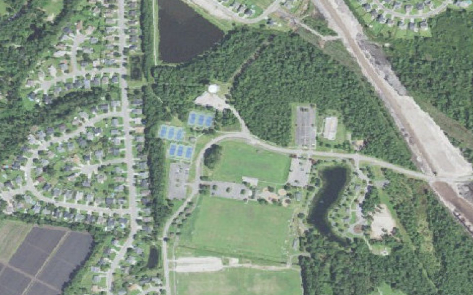 Aerial view of Ogden Park in Ogden, North Carolina, showing its tennis courts, open playing fields, parking and pond, with the Military Cutoff Road extension corridor under construction at right