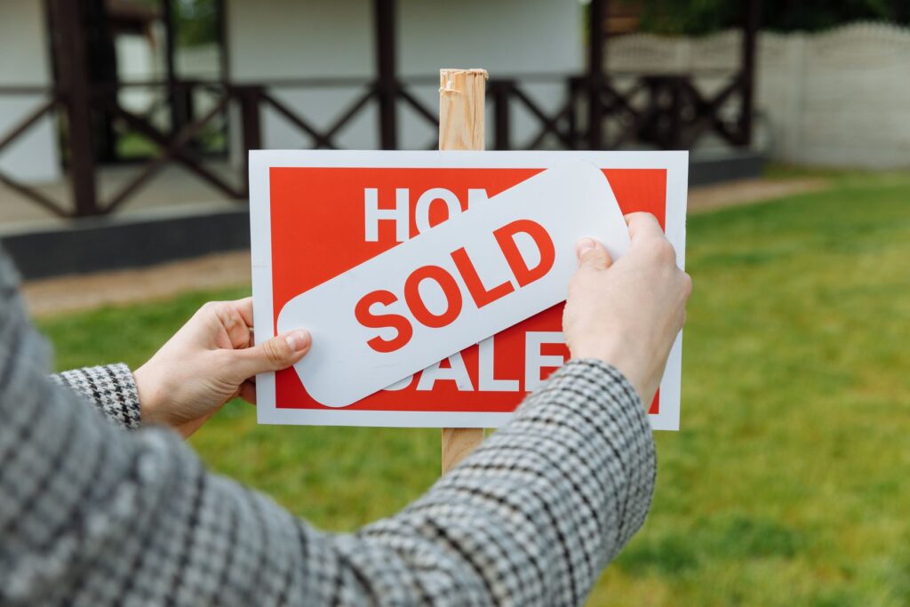 A hand pressing a SOLD sticker onto a red and white real estate For Sale sign in a front yard