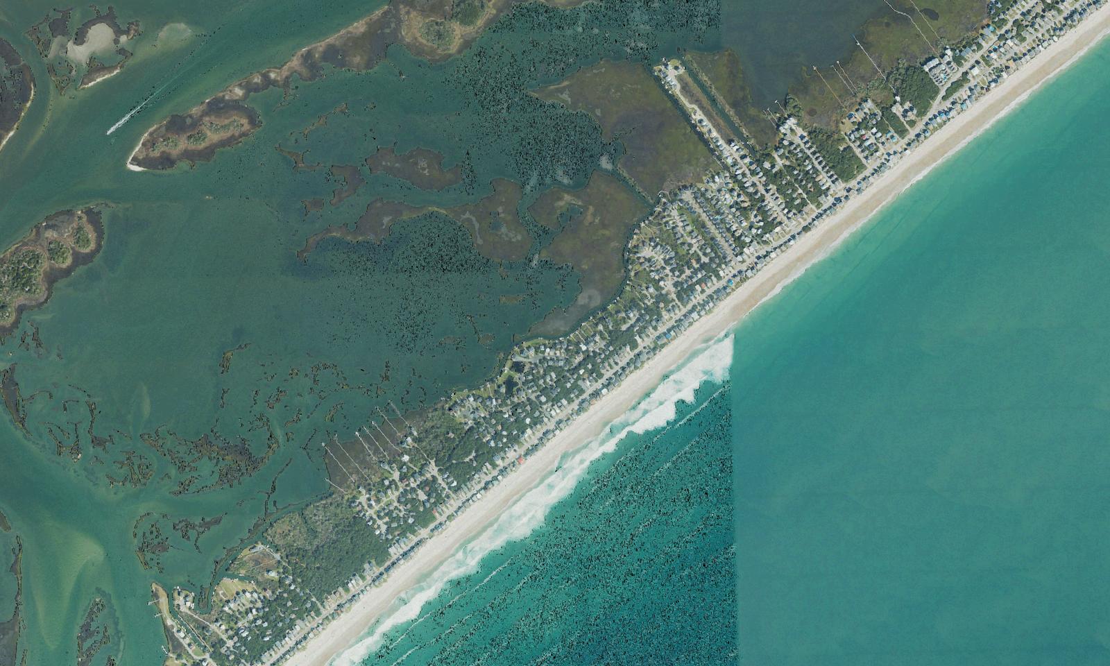 Aerial view of the south end of Surf City, North Carolina, with a narrow single row of beachfront houses, docks and piers reaching into the sound, wide marsh and tidal flats behind the island and the Intracoastal Waterway beyond
