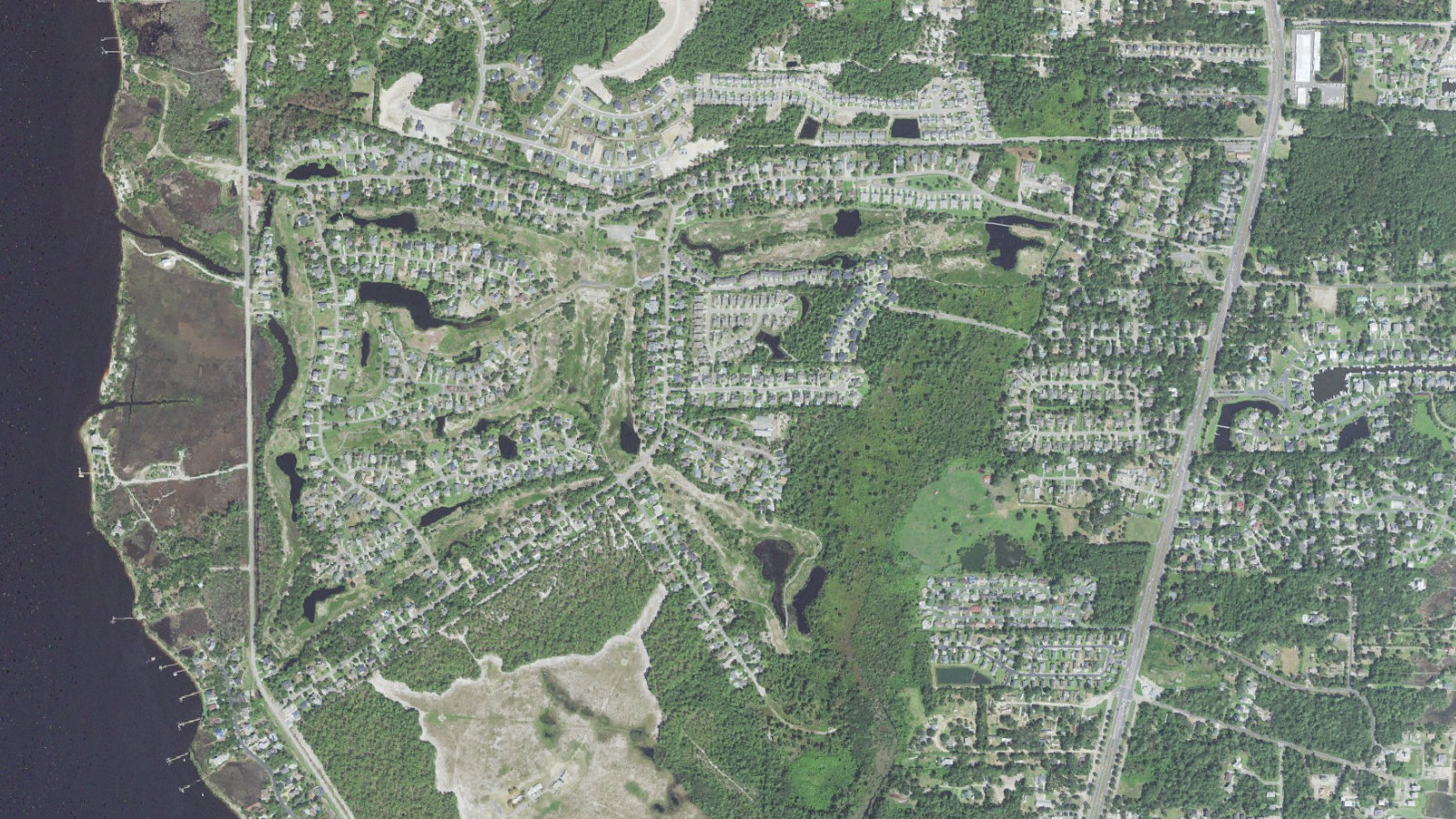 Aerial view of The Cape in unincorporated New Hanover County, North Carolina, with the Cape Fear River and River Road along the left edge, curving streets and lagoons through the center, the wide bare corridors of the closed golf course winding between them, and Carolina Beach Road at right