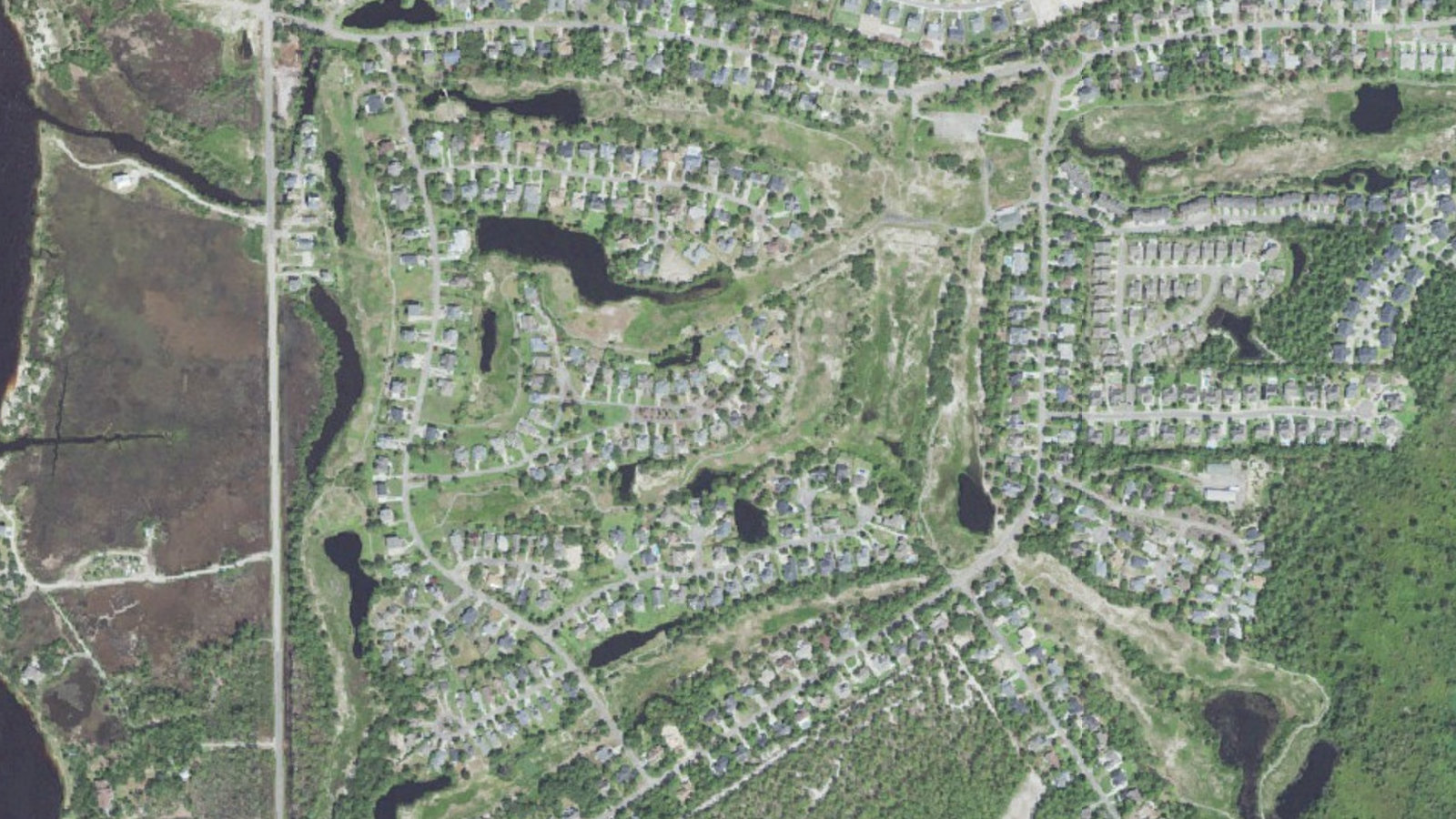 Closer aerial view of the middle of The Cape, Wilmington, North Carolina, showing houses on curving streets around stormwater lagoons, the unmaintained fairway corridors of the former Cape Golf and Racquet Club running through the neighborhood, and the marsh and Cape Fear River at left