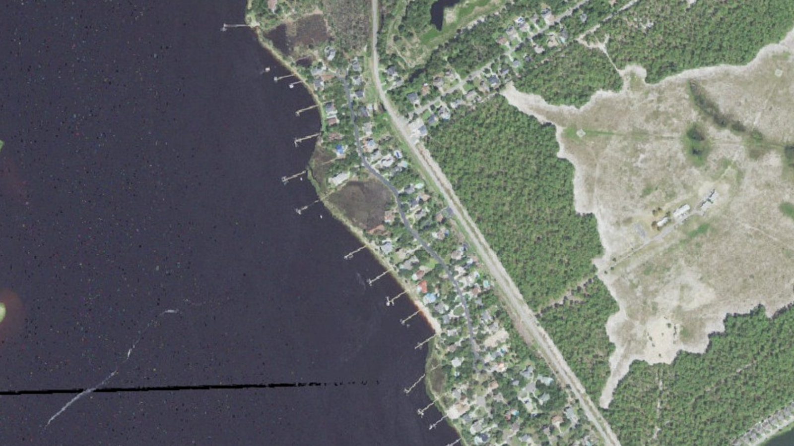 Aerial view of the riverfront section of The Cape, Wilmington, North Carolina, with Shipwatch homes between River Road and the Cape Fear River and their long private piers reaching across the shallows