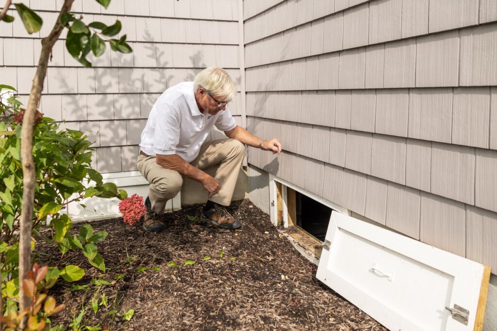 A person kneeling to inspect a crawl space entrance at the side of a house