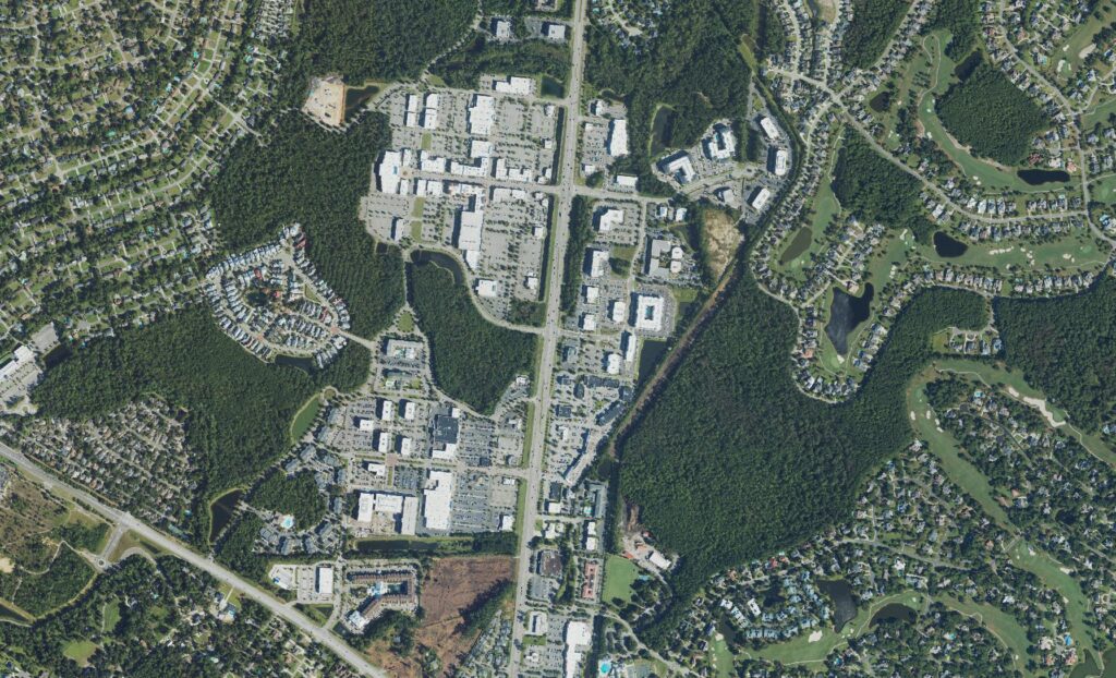 Aerial view of the Military Cutoff Road corridor in midtown Wilmington, North Carolina, showing the Mayfaire shopping district and its parking fields at center, surrounding apartment and single-family subdivisions under heavy pine canopy, and the golf course fairways and ponds of the Landfall community along the eastern edge