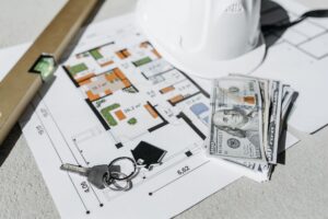 A floor plan on a table with cash, house keys and a hard hat, representing the money side of a home purchase