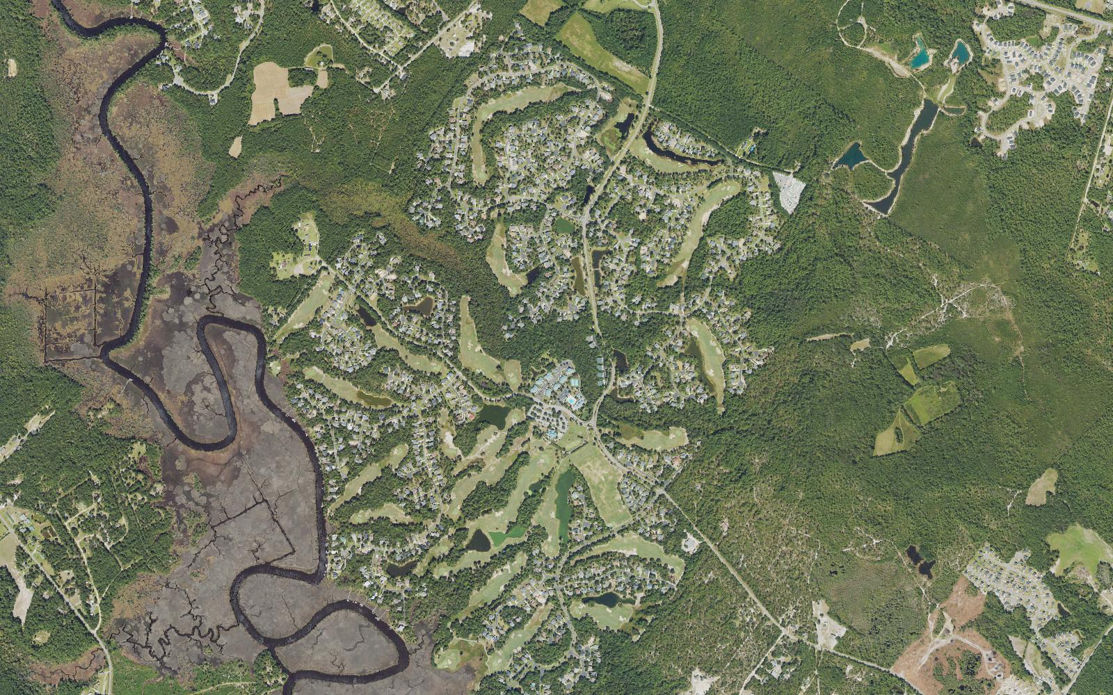 Aerial view of Winding River Plantation in Bolivia, North Carolina, showing the Carolina National golf fairways threaded between wooded homesites and ponds, with the Lockwood Folly River and its salt marsh along the western edge